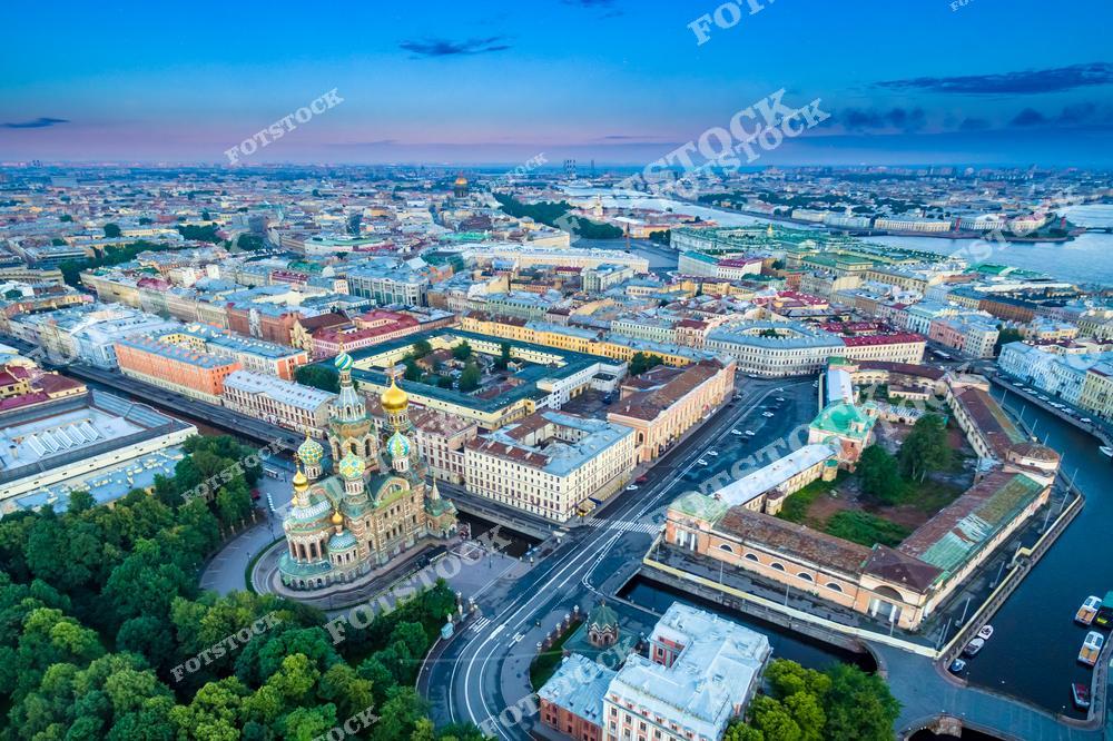 Панорамная фотография Санкт-Петербурга с высоты птичьего полета на летнем закате. На снимке видны улицы города, мосты через Неву, река и уникальная архитектура исторического центра. Съемка с дрона позволяет показать масштабы застройки, панораму набережных и красивые отражения солнечного света в воде. Фотография передает атмосферу летнего Петербурга, его историческое и культурное наследие, а также гармонию между городской архитектурой и природными элементами. Снимок подходит для туристических публикаций, фотостоков, оформления интерьеров, рекламы и материалов о путешествиях по Северной столице России.